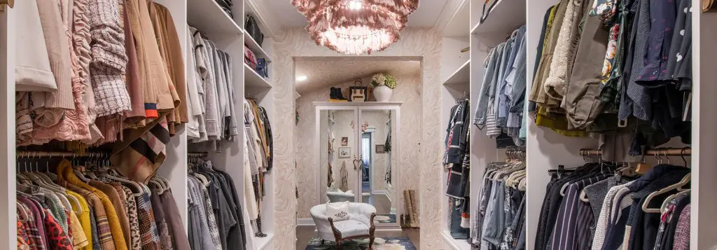 A stylish walk-in closet with built-in shelves and hanging racks, showcasing a variety of colorful clothing, a mirrored cabinet at the far end, elegant wallpaper