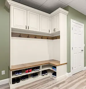 Mudroom storage unit with white cabinetry, upper cabinets, wooden bench seating, hooks for hanging, and cubbies neatly organized with shoes against a green wall