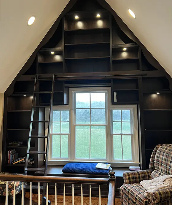 Custom built-in bookcase with a rolling ladder and integrated lighting, surrounding a large window with a cushioned bench seat, set in a vaulted ceiling room with dark wood cabinetry and cozy reading area