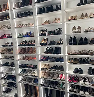 Custom-built white shelving system displaying a large, organized collection of women’s shoes, ranging from boots to heels to sneakers, all neatly lit with integrated LED lighting