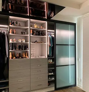 Contemporary custom closet with built-in drawers, illuminated shelving for shoes and accessories, and frosted glass sliding doors