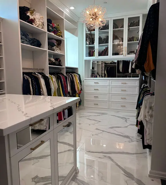 Luxury white walk-in closet featuring custom cabinetry, glass-front upper cabinets, spacious drawers, hanging sections, a display island, crystal chandelier, and polished marble-look flooring.