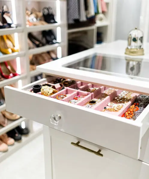 Custom jewelry drawer with pink velvet inserts and crystal hardware in a white walk-in closet, featuring a glass-top island and lighted shoe rack storage.