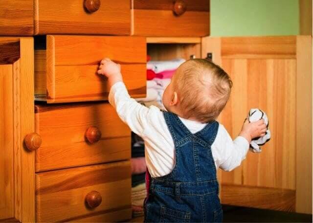 An infant in overalls opens a wooden drawer while holding a napkin 