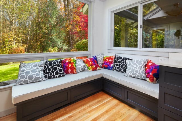 A collection of vibrant pillows sit on a corner bench in an enclosed porch