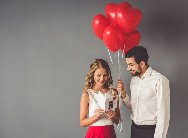 Two young people. One has given the other a valentines card while holding a bunch of heart shaped balloons