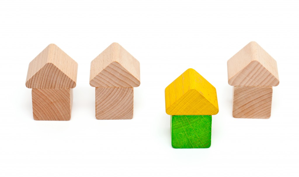 Four toy wooden houses in a row. three are unpainted. One is painted yellow and green and stands in front of the others