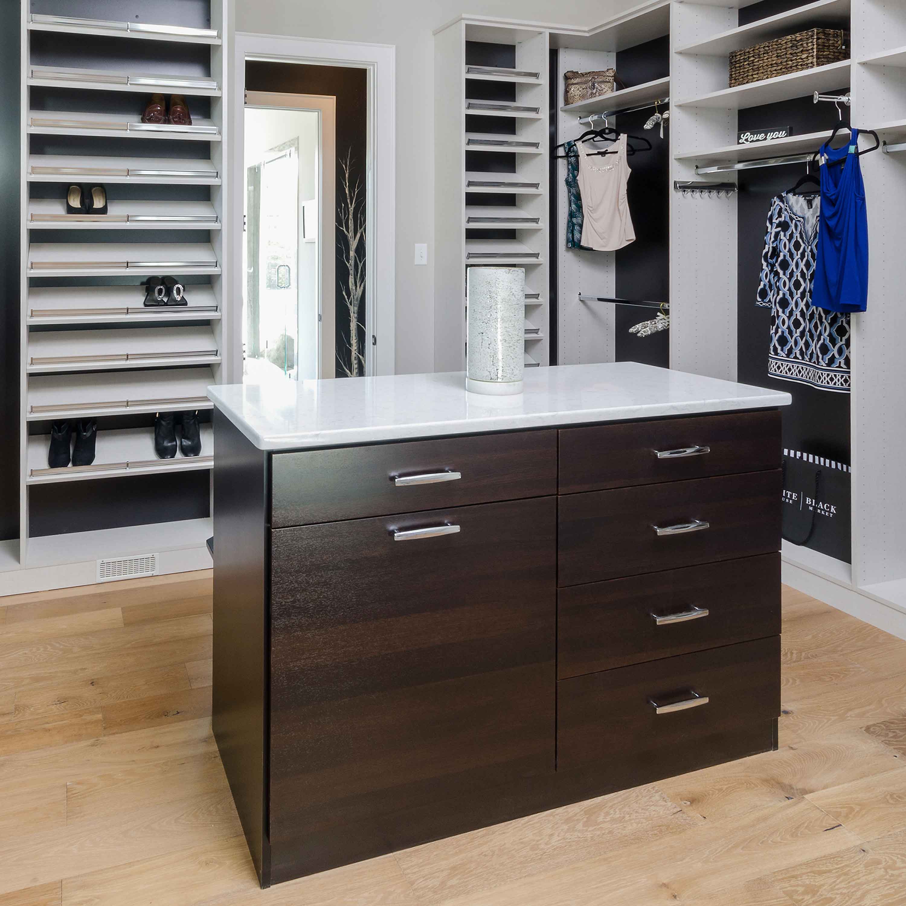 A dark wood island with a white marble top sits in the center of a white closet