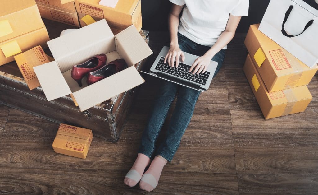 Woman sits on the floor on her laptop. She is surrounded by clothes she is listing for sale