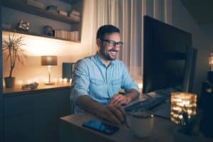 A man smiles while working on is computer in a dimly lit office