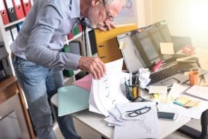 A cluttered desk with a man searching for something under a stack of paper