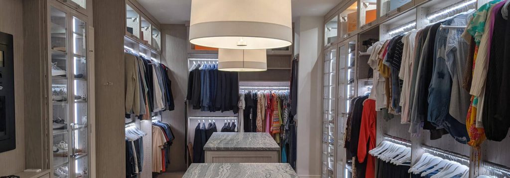 Spacious custom walk-in closet featuring dual islands with stone countertops, illuminated hanging sections, glass-front cabinetry, and modern pendant lighting