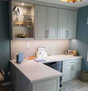Custom-built home office featuring an L-shaped white desk with cabinetry above and below, set against a textured backsplash and soft blue walls