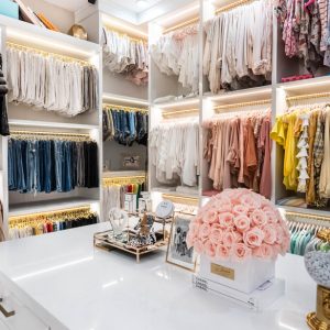 Custom white melamine system with triple-hang wardrobe rods, gold-tone hanging hardware, and integrated LED lighting. Central island provides additional accessory organization space.