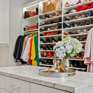 Custom closet system in White Chocolate Melamine with full-height shoe shelving, double-hang wardrobe rods, and a marble-look island. Open cubbies and integrated lighting highlight accessories and footwear.