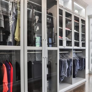 Glass-front wardrobe system in Dove Grey with integrated LED lighting, double-hang rods, and display shelving. Features stacked drawer banks, acrylic shelf dividers, and a mirrored vanity station.