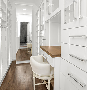 Bright, elegant walk-in closet with white cabinetry, glass drawer pulls, a built-in vanity, and a classic upholstered chair on dark hardwood floors