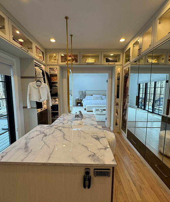 Luxurious walk-in closet featuring a marble island, mirrored cabinetry, upper display shelves, and a view into a bright, modern bedroom
