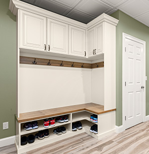 Mudroom storage unit with white cabinetry, upper cabinets, wooden bench seating, hooks for hanging, and cubbies neatly organized with shoes against a green wall