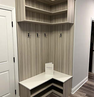 Corner mudroom unit featuring upper open shelving, four metal hooks, and a white bench with under-seat cubbies, designed in a light wood grain finish
