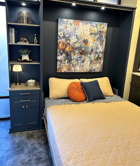 Navy blue wall bed with built-in shelving and drawers, featuring a cozy beige quilt, colorful accent pillows, and a floral painting above the headboard