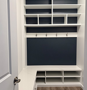 Custom-built white mudroom with navy blue back panels, open cubbies for shoes, upper shelving for storage, and wall hooks for hanging items