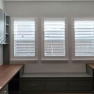Custom home office with two built-in desks, upper cabinets, open shelving, storage drawers, and three large windows with plantation shutters.