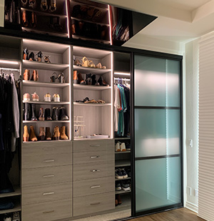 Contemporary custom closet with built-in drawers, illuminated shelving for shoes and accessories, and frosted glass sliding doors