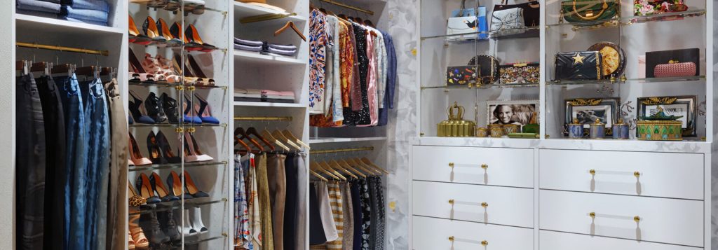 Elegant walk-in closet featuring white cabinetry, glass shelving with designer handbags, gold accents, and organized hanging sections for pants, skirts, and jackets