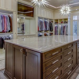Spacious closet with cream-toned cabinets, a large island with deep drawer storage, built-in glass shoe shelving, and double hanging rods.
