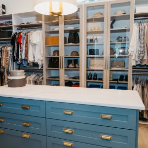 Walk-in closet with a bold blue island featuring framed drawer fronts and brass hardware