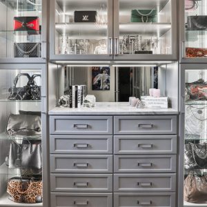 Modern gray closet with integrated lighting, glass display shelves for designer handbags, and a dedicated vanity station with full-extension drawers.