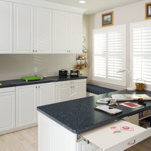 Custom craft room Raised Panel cabinet doors in Antique White melamine with a dark solid-surface countertop. Features a slide-out ironing drawer, lower open shelving, and wraparound work surface.