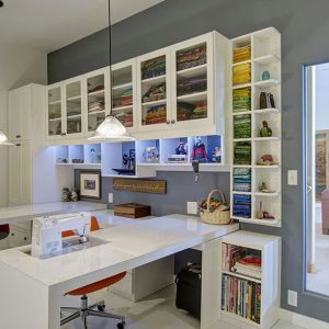 Craft room organization with Glass-front upper cabinets with Shaker frames in White Chocolate melamine. Includes vertical fabric cubbies, open book shelving, and a built-in sewing workstation with wraparound desk.