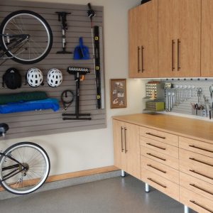 Warm wood-toned garage storage with bikes mounted on slat wall panels and cabinetry organizing helmets, tools, and accessories.