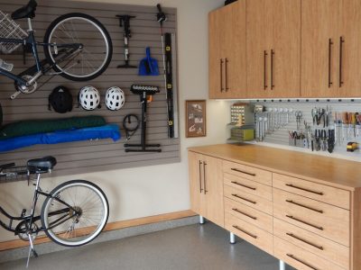 Warm wood-toned garage storage with bikes mounted on slat wall panels and cabinetry organizing helmets, tools, and accessories.