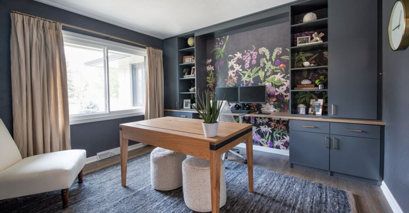 Modern home office with floral accent wallpaper, matte charcoal built-ins, and a light oak desk flanked by ottomans, offering ample shelf space