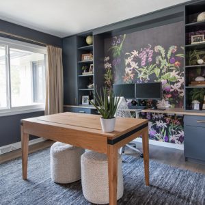 Modern home office with floral accent wallpaper, matte charcoal built-ins, and a light oak desk flanked by ottomans, offering ample shelf space
