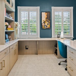 Light wood-finish home office cabinetry surrounds an L-shaped quartz desk, featuring built-in bookshelves