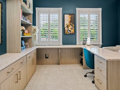 Light wood-finish home office cabinetry surrounds an L-shaped quartz desk, featuring built-in bookshelves