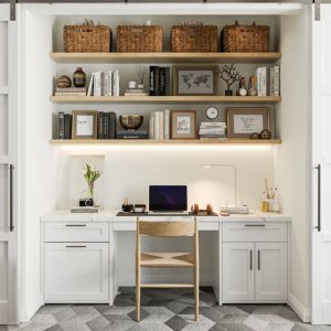 Compact, closet-style workstation with two-tier open shelving, wicker baskets, framed prints, and a flush-mount LED task light