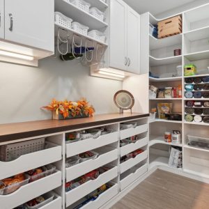 Custom white pantry with multiple open pull-out drawers and upper cabinetry, featuring a wood slab countertop and wine glass holders.