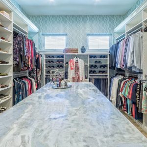 Large central island and dual handing rods in walk-in closet. Features glass open shelving and crown molding.