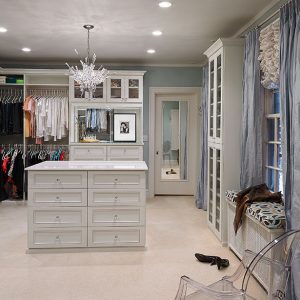 Expansive dressing room featuring a central island with drawer storage, dual-level hanging rods, integrated wardrobe cabinetry with glass doors, and upper storage